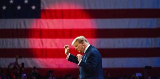 Man in suit gesturing on stage with flag background.