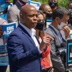 Man speaking at campaign event with supporters behind him.
