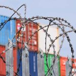 Barbed wire fence with shipping containers in background.