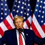 Man speaking on stage with multiple American flags