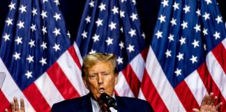Man speaking on stage with multiple American flags