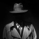 Person in trench coat and fedora face obscured by shadow