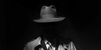 Person in trench coat and fedora face obscured by shadow