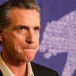 California’s Election Bombshell: Power Shift Looms Man in suit, solemn expression, purple background with bear drawing.