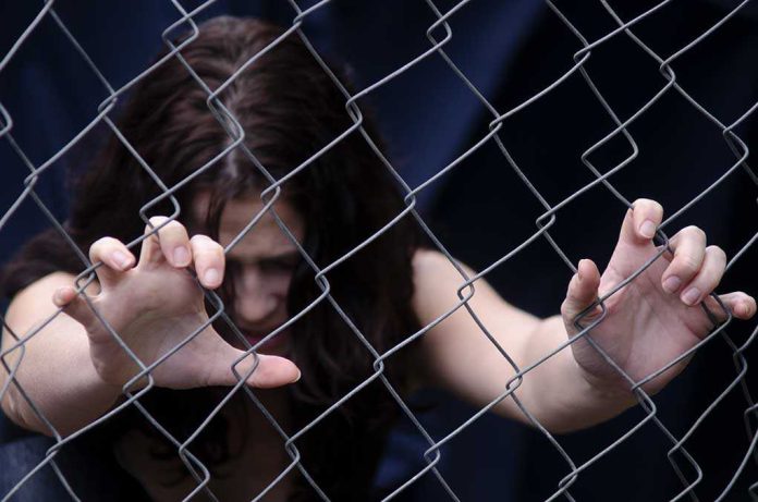 A person with long dark hair reaching through a metal fence, expressing distress