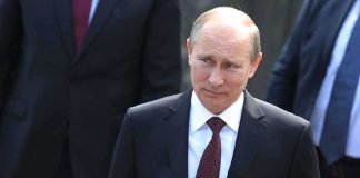 A serious-looking man in a suit walking outdoors with security personnel in the background