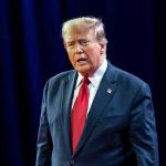 Trump Accused of Racial Profiling — Outrage Erupts! Man in suit with red tie speaking on stage.