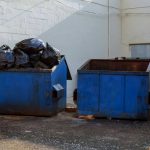 Two blue dumpsters, one overflowing with trash bags.