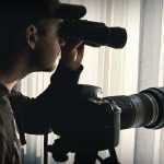 Millionaires Minted From ABSURD Surveilance Program Person looking through binoculars beside a camera.
