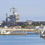 A large naval aircraft carrier docked in a harbor with smaller boats in the foreground