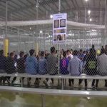 People sitting on benches inside a fenced facility.