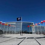 Putin Declares Ukraine War ‘Ending’ – Real or Ruse? Flags outside NATO headquarters building under clear blue sky.