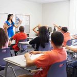 Demographic Earthquake Hits Classrooms Teacher in a blue dress instructing students in a classroom with hands raised