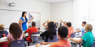 Demographic Earthquake Hits Classrooms Teacher in a blue dress instructing students in a classroom with hands raised