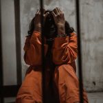 Person in orange jumpsuit sitting behind bars, handcuffed.