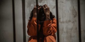 Budget Bombshell Freezes Trans Care Person in orange jumpsuit sitting behind bars, handcuffed.
