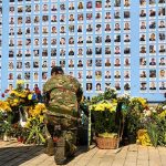 Soldier kneels before memorial wall with photos and flowers