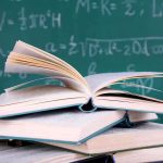 A stack of books with one open on top, in front of a chalkboard filled with mathematical equations