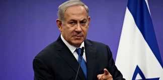 man in suit speaking at podium with Israeli flag
