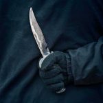 Gloved hand holding a serrated knife against a dark jacket