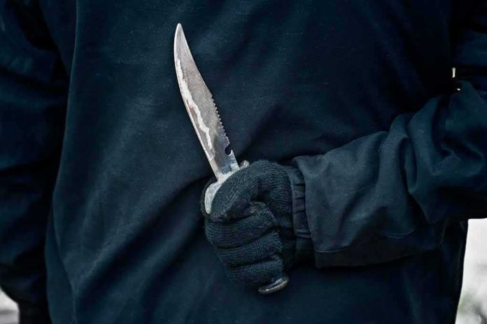 shutterstock_1373715260 Gloved hand holding a serrated knife against a dark jacket