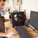 Man operating a 3D printer next to a computer with CAD designs