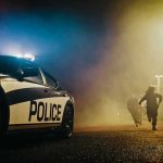 Police car with flashing lights at night as two people run in a foggy alley