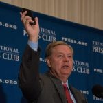 Bipartisan Power Play After Lindsey Graham’s Shocking Death Man in suit speaking at podium holding up a smartphone