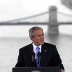 25 Years Later: Vigilance Or Complacency? A man in a suit giving a speech at a podium with a bridge in the background