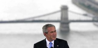 25 Years Later: Vigilance Or Complacency? A man in a suit giving a speech at a podium with a bridge in the background