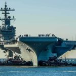 San Diego Cheers, Silent Crisis Lurks Aircraft carrier guided by tugboats in a harbor