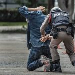 Ex-Spy Boss Nabbed – Treason Bombshell Police officer handcuffing person kneeling on pavement