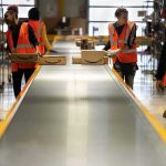 Warehouse workers in orange vests handle packages on a conveyor belt