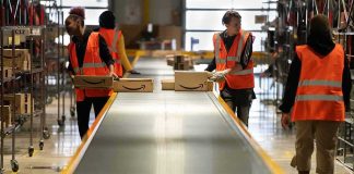 Warehouse workers in orange vests handle packages on a conveyor belt