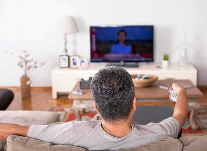 shutterstock_1535770295 Man watching TV from couch with remote in hand