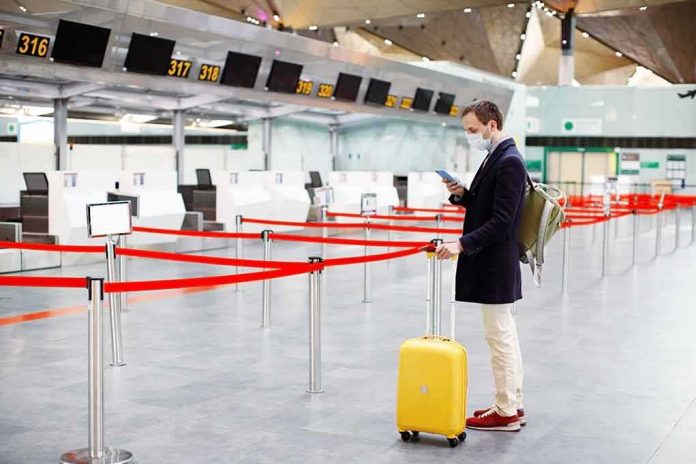 shutterstock_1684988143-1 Traveler wearing a mask stands by yellow suitcase at empty airport check-in